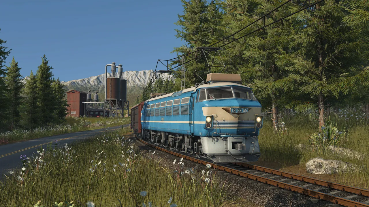 A blue and cream electric locomotive hauling freight wagons past pine trees and a silo plant, snowy peaks behind