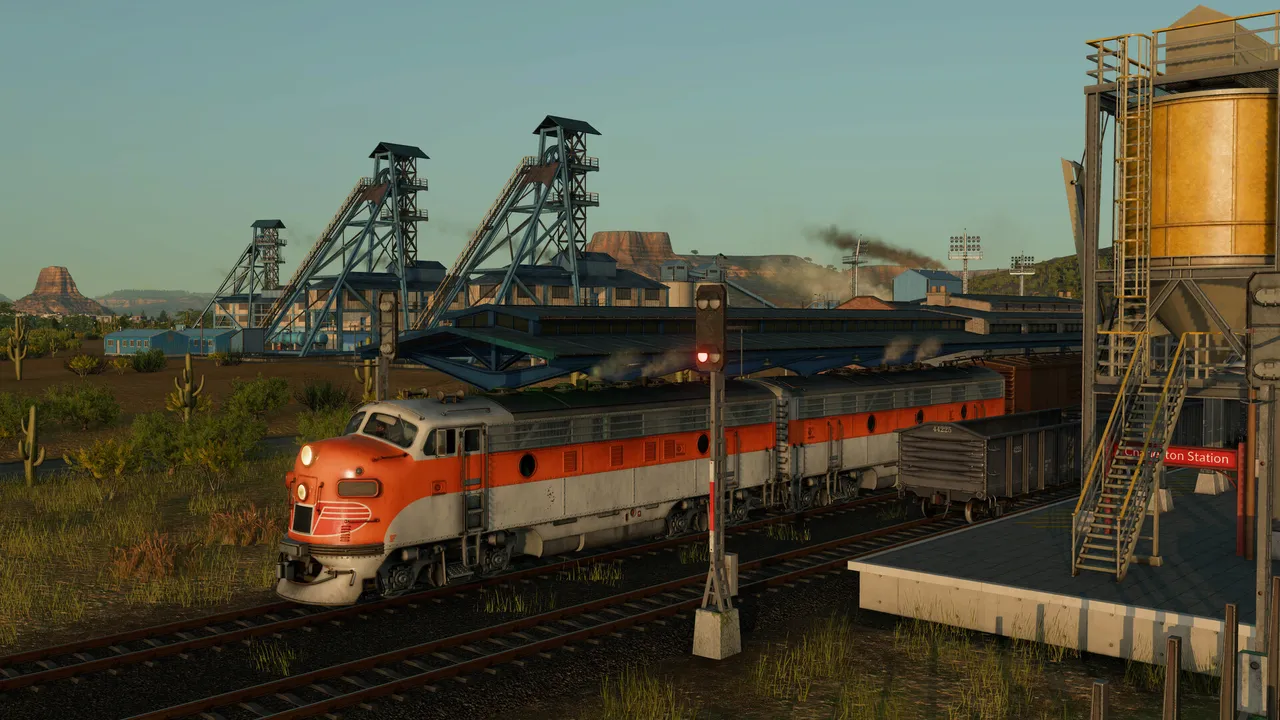 An orange and silver diesel locomotive at a station platform with a red signal lamp, mine headframes behind